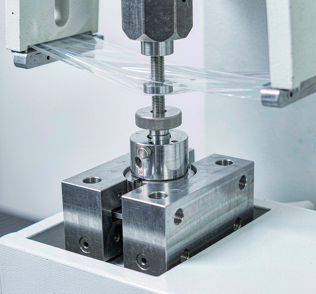 Laboratory tensile testing machine gripping a strip of clear plastic film during a strength test