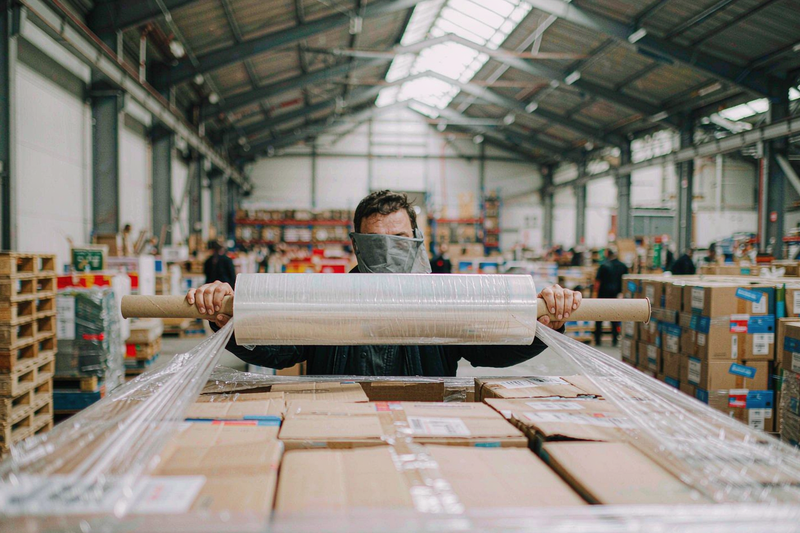 Warehouse worker holding an extended core hand stretch film roll with both hands, cardboard handles visible beyond the film